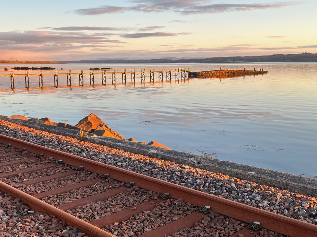 Beach at Culross, looking over the water with train tracks in the foreground and a pier in the background.