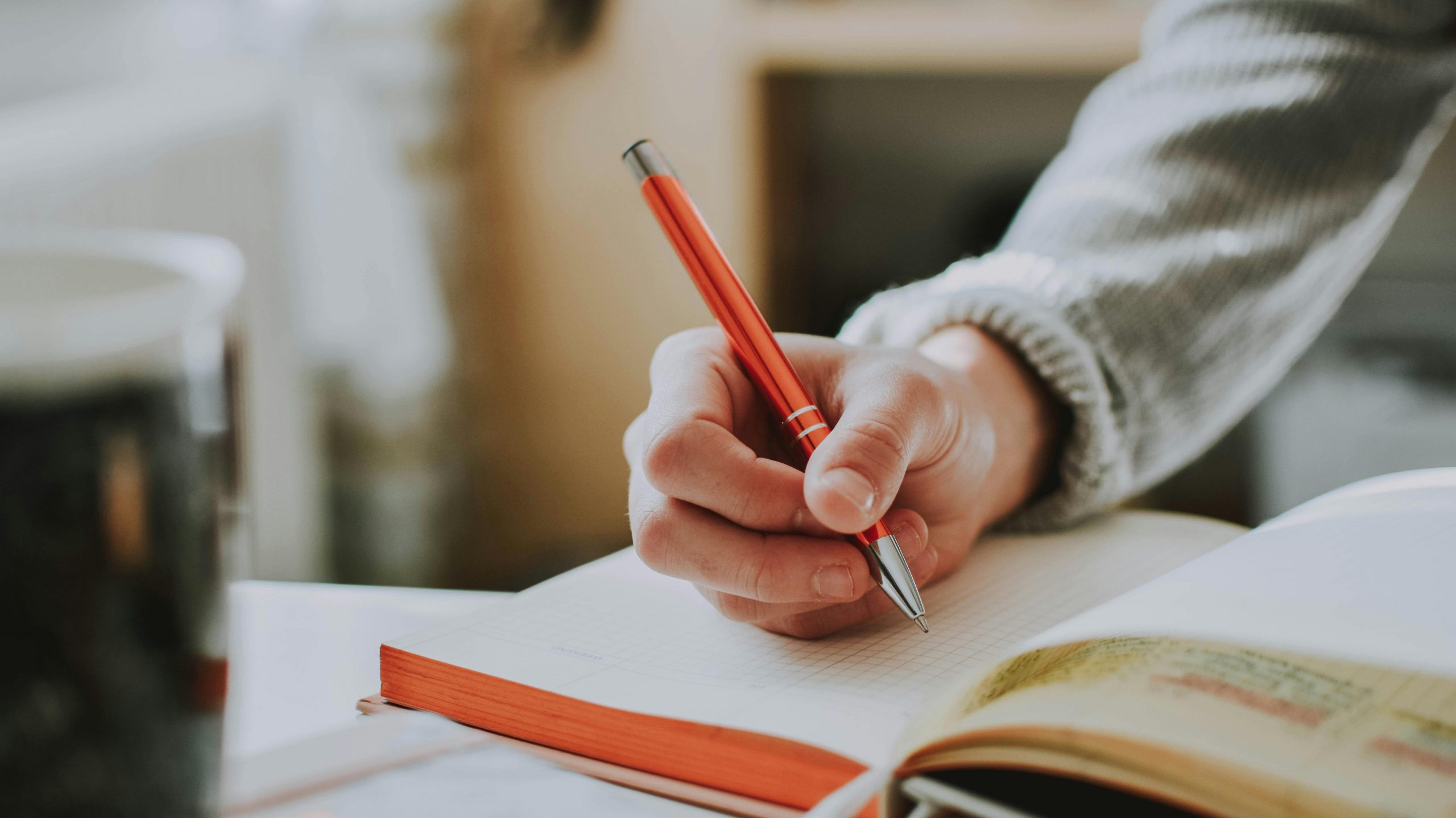 You only see the arm, dressed in a grey sweatshirt, holding a pen, about to write in a notebook that sits on a plain white desk.