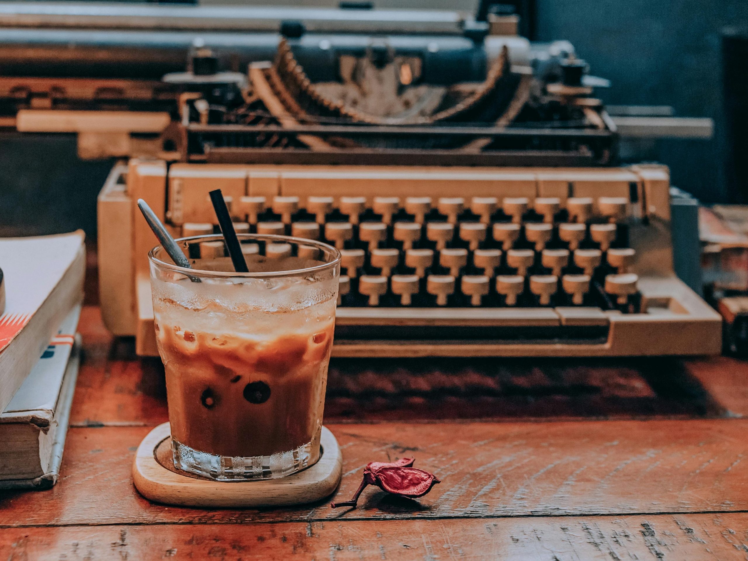 A typewriter on a rustic wooden desk with notebooks on the left hand side an glass of ice coffe in front beside a dried rosebud 